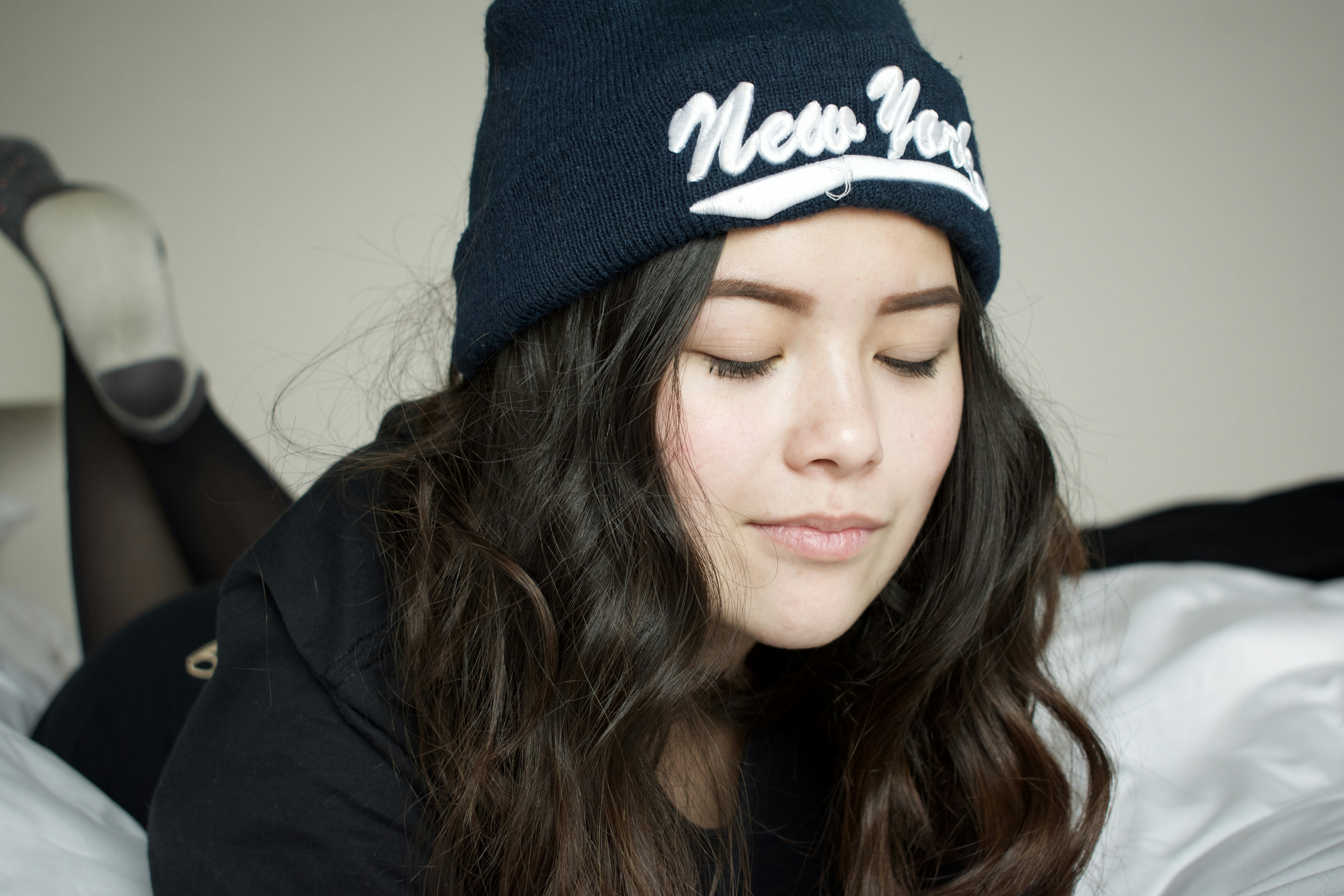 woman lying on white bed wearing black New York knit cap