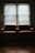 Two armchairs with brown cushions sit side by side in front of a window covered by blinds. The room is dimly lit with subdued natural light filtering through the blinds, casting shadows and creating a serene, quiet atmosphere. The wooden floor adds warmth to the space.