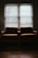 Two armchairs with brown cushions sit side by side in front of a window covered by blinds. The room is dimly lit with subdued natural light filtering through the blinds, casting shadows and creating a serene, quiet atmosphere. The wooden floor adds warmth to the space.