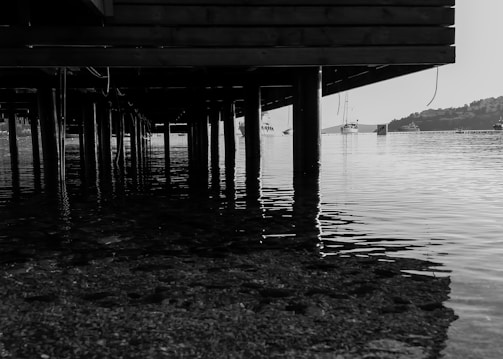 A black and white photograph taken from beneath a wooden pier. The wooden structure is supported by multiple vertical pillars that extend into the calm, reflective water. In the background, there are a few boats docked or anchored on the water. The shoreline is visible in the distance with some buildings and trees on a hillside.