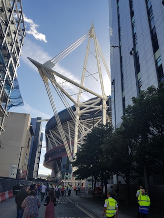 A large sports stadium with towering white architectural structures is framed by modern buildings on either side. People are gathered on the street, some wearing high-visibility vests, possibly staff or security. Trees line one side of the street, and the sky is clear with a few clouds.