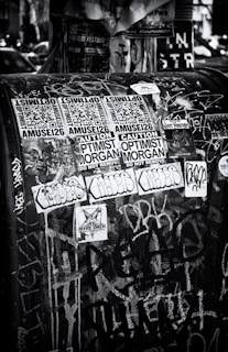 A black and white urban scene featuring a public space covered with various stickers and graffiti. The stickers include QR codes, warning labels, and artistic designs, layered over a backdrop of chaotic spray paint and marker scribblings.