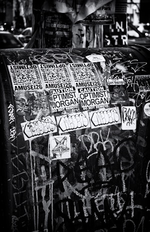 A black and white urban scene featuring a public space covered with various stickers and graffiti. The stickers include QR codes, warning labels, and artistic designs, layered over a backdrop of chaotic spray paint and marker scribblings.