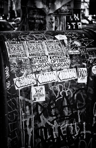 A black and white urban scene featuring a public space covered with various stickers and graffiti. The stickers include QR codes, warning labels, and artistic designs, layered over a backdrop of chaotic spray paint and marker scribblings.