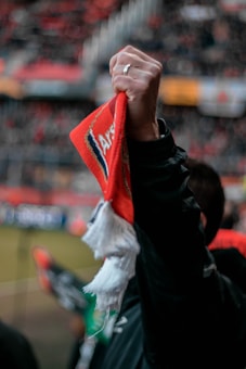 A close-up of a hand holding a red and white sports scarf aloft at a stadium. The blurred background suggests a crowd and stadium seating, indicating a sporting event atmosphere.