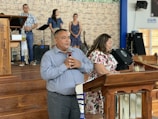 A man and a woman stand at a wooden pulpit in a church setting. The man holds a microphone, suggesting he might be speaking or addressing an audience. Behind them, a group of people are positioned on a stage with musical equipment, including a music stand and a guitar. The background features a brick wall, and there's a sign on the wall that says 'Salida.'