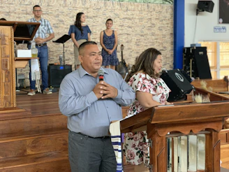 A man and a woman stand at a wooden pulpit in a church setting. The man holds a microphone, suggesting he might be speaking or addressing an audience. Behind them, a group of people are positioned on a stage with musical equipment, including a music stand and a guitar. The background features a brick wall, and there's a sign on the wall that says 'Salida.'