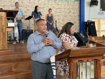 A man and a woman stand at a wooden pulpit in a church setting. The man holds a microphone, suggesting he might be speaking or addressing an audience. Behind them, a group of people are positioned on a stage with musical equipment, including a music stand and a guitar. The background features a brick wall, and there's a sign on the wall that says 'Salida.'