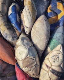 A collection of old, deflated sports balls, including footballs and volleyballs, arranged closely together. The balls display signs of wear, with faded colors and frayed surfaces. Text from brands such as 'Cosco' and 'Vega' is visible on some of them.
