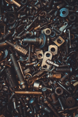 A chaotic assortment of various metallic bolts, nuts, washers, and screws. The parts are densely packed, creating a textured and industrially themed image. The lighting highlights some metal pieces with a warm glow, adding depth and contrast.