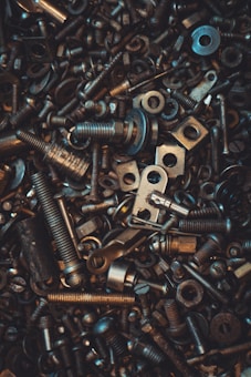 A chaotic assortment of various metallic bolts, nuts, washers, and screws. The parts are densely packed, creating a textured and industrially themed image. The lighting highlights some metal pieces with a warm glow, adding depth and contrast.