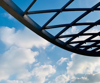 A metal structure with a grid-like pattern is silhouetted against a backdrop of bright blue sky and fluffy white clouds. The structure appears to be part of a roof or canopy, and the scene conveys a sense of openness and space.