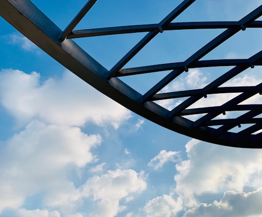 A metal structure with a grid-like pattern is silhouetted against a backdrop of bright blue sky and fluffy white clouds. The structure appears to be part of a roof or canopy, and the scene conveys a sense of openness and space.