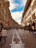 A bustling street scene in a European city with historic architecture lining both sides. People are walking along the cobblestone street and tram tracks run down the center. A Swiss flag is prominently displayed on one of the buildings.