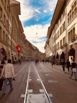A bustling street scene in a European city with historic architecture lining both sides. People are walking along the cobblestone street and tram tracks run down the center. A Swiss flag is prominently displayed on one of the buildings.