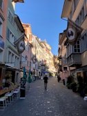 A narrow, cobblestone street is lined with colorful buildings featuring various architectural designs. Flags hang from the upper floors, advertising a film festival. Outdoor seating and small shops are visible along the street. People are casually strolling, enjoying the sunny weather.