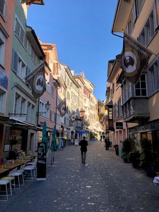 A narrow, cobblestone street is lined with colorful buildings featuring various architectural designs. Flags hang from the upper floors, advertising a film festival. Outdoor seating and small shops are visible along the street. People are casually strolling, enjoying the sunny weather.