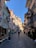 A narrow, cobblestone street is lined with colorful buildings featuring various architectural designs. Flags hang from the upper floors, advertising a film festival. Outdoor seating and small shops are visible along the street. People are casually strolling, enjoying the sunny weather.