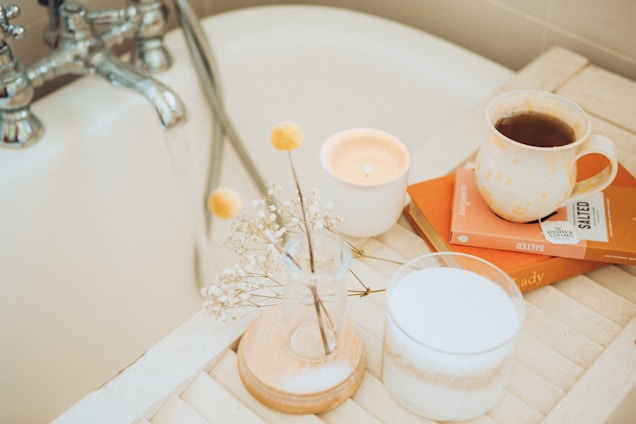 A cozy bathroom scene featuring a white bathtub with chrome faucets. A wooden tray rests across the tub, holding an arrangement of delicate flowers in a small glass vase, a lit candle, a white ceramic mug filled with coffee, and two books with orange covers.