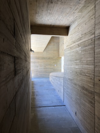 A minimalist interior space featuring a long, narrow hallway constructed with smooth, textured concrete walls and ceiling. The floor is made of light tiles, and the space is illuminated by natural light entering from an unseen window at the end of the corridor.
