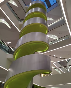A modern interior structure featuring a large spiral staircase with bright green accents and sleek gray panels. The architecture displays clean lines and geometric forms, with multiple levels visible. Overhead lighting casts a neutral glow throughout the space.