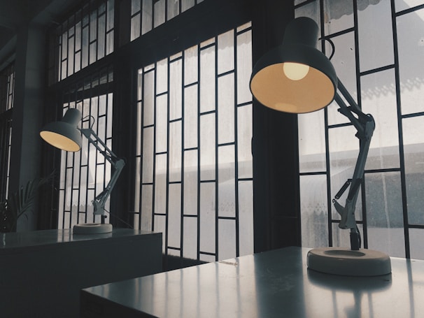 Two angled desk lamps sit on a reflective surface near a large window with a grid-like frame. The light from the lamps creates a warm glow in contrast to the cool, muted light coming through the window, casting shadows on the surface.
