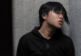 A young person with medium-length dark hair leans against a textured gray wall with eyes closed and an expression suggesting emotional distress or fatigue. They are wearing a black jacket with a hood.