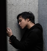 A person in a black hoodie is leaning against a rough, textured wall, looking thoughtfully or pensively at an object in their hand.