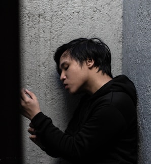 A person in a black hoodie is leaning against a rough, textured wall, looking thoughtfully or pensively at an object in their hand.
