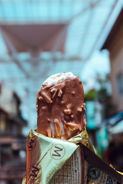 A close-up of an ice cream bar with a chocolate coating and almond pieces, partially unwrapped in a gold and brown wrapper. The background is blurred, showing the outlines of a street or market with overhead structures.