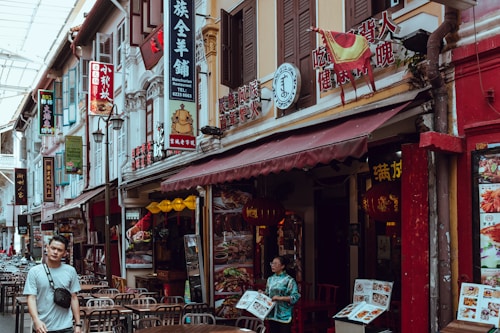 A busy street in an Asian city lined with various shops and restaurants. Numerous signs with Chinese characters and vibrant colors adorn the buildings. Outdoor tables are set, suggesting dining establishments. At least two people are visible, one walking and another holding a menu outside a restaurant.