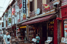 A busy street in an Asian city lined with various shops and restaurants. Numerous signs with Chinese characters and vibrant colors adorn the buildings. Outdoor tables are set, suggesting dining establishments. At least two people are visible, one walking and another holding a menu outside a restaurant.