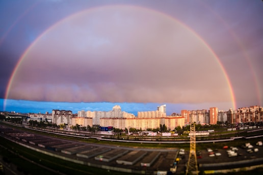 A vibrant cityscape features a prominent double rainbow arching over a cluster of high-rise buildings. The sky transitions from bright, sunlit clouds to a darker, stormy hue. Below, a mix of roads and train tracks is visible, with various vehicles and buildings in the foreground.
