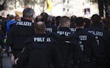 A group of uniformed police officers, with 'Polizei' written on their backs, are gathered, possibly at a public event or protest. They are in a close formation, suggesting they are on duty and maintaining order. Various signs and a crowd are visible in the background.