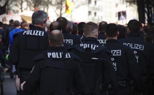A group of uniformed police officers, with 'Polizei' written on their backs, are gathered, possibly at a public event or protest. They are in a close formation, suggesting they are on duty and maintaining order. Various signs and a crowd are visible in the background.