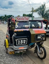 A colorful motorized rickshaw with a 'Special Trip' sign is parked on the side of a paved road. The vehicle has a black and white number plate reading '157'. It appears slightly worn and has various stickers on its body. The background includes several palm trees, electrical wires, and a partly cloudy sky with other vehicles visible on the road.