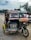 A colorful motorized rickshaw with a 'Special Trip' sign is parked on the side of a paved road. The vehicle has a black and white number plate reading '157'. It appears slightly worn and has various stickers on its body. The background includes several palm trees, electrical wires, and a partly cloudy sky with other vehicles visible on the road.
