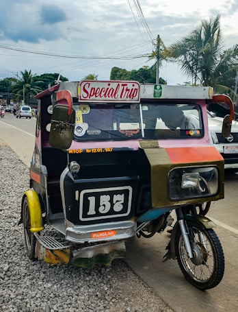 A colorful motorized rickshaw with a 'Special Trip' sign is parked on the side of a paved road. The vehicle has a black and white number plate reading '157'. It appears slightly worn and has various stickers on its body. The background includes several palm trees, electrical wires, and a partly cloudy sky with other vehicles visible on the road.