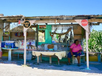 A small outdoor market stall with a corrugated metal roof and wooden supports. Various handmade crafts and decorations hang from the structure, with a table displaying items for sale. There are colorful bottles on a makeshift table to the left. A person wearing a pink shirt is seated on the right side, under a partial shade, looking to the left. Signs reading 'Follow Your Heart' and 'Coca-Cola' are visible.