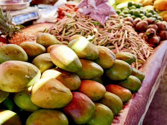 A market stall displaying an abundance of fresh, ripe mangoes piled on top of each other, with various other produce items like beans and pineapples visible in the background, all under a warm and natural light.