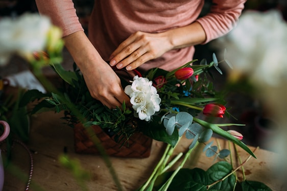 Hands are arranging a floral bouquet with white and pink flowers amidst various green leaves, set on a wooden table. The person appears to be wearing a long-sleeved, light pink top.