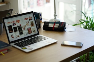 A workspace featuring a laptop displaying a variety of images, next to a pencil case with pens, a smartphone, and a book. The setting includes a potted plant and desk organizer, suggesting a creative and organized environment.