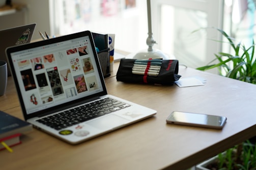 A workspace featuring a laptop displaying a variety of images, next to a pencil case with pens, a smartphone, and a book. The setting includes a potted plant and desk organizer, suggesting a creative and organized environment.