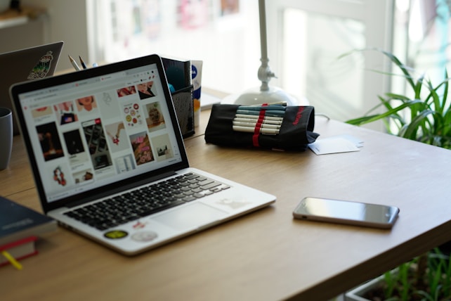 A workspace featuring a laptop displaying a variety of images, next to a pencil case with pens, a smartphone, and a book. The setting includes a potted plant and desk organizer, suggesting a creative and organized environment.