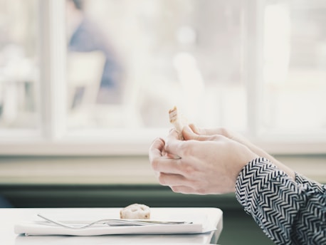 A person wearing a patterned long-sleeve shirt holds a small piece of food in their hands. The setting appears to be indoors, with a window in the background allowing in soft natural light. A table is partially visible, with a plate and a fork resting on a folded napkin.