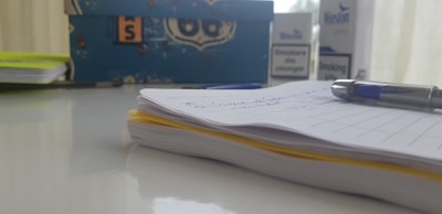 A close-up of a notebook with handwritten notes, alongside a blue pen resting on it. In the background, there is a decorative box and a box of cigarettes. The setting appears to be on a reflective white surface, likely a desk.