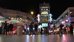 A busy nightlife scene with neon signs and illuminated advertisements. People are gathered and walking around a vibrant street, with various buildings and signs showcasing businesses like Slotzilla, Walgreens, and others. The atmosphere is lively with bright lights and crowds enjoying the night.