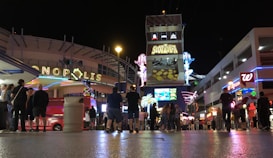 A busy nightlife scene with neon signs and illuminated advertisements. People are gathered and walking around a vibrant street, with various buildings and signs showcasing businesses like Slotzilla, Walgreens, and others. The atmosphere is lively with bright lights and crowds enjoying the night.