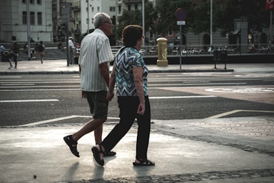 An elderly couple walks hand in hand across a city street. The urban environment features buildings, parked bicycles, and blurred figures in the background. The scene captures a moment of quiet companionship amidst a busy setting.