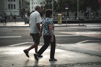 An elderly couple walks hand in hand across a city street. The urban environment features buildings, parked bicycles, and blurred figures in the background. The scene captures a moment of quiet companionship amidst a busy setting.